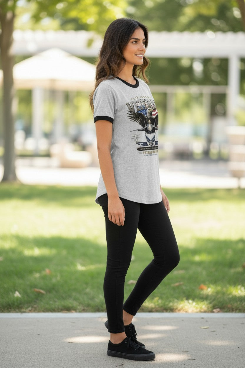 A woman modeling a light gray ringer t-shirt with black trim and a graphic print of an eagle and the words 'WILD WEST', paired with black leggings and black sneakers, standing outdoors on a paved path with a blurred park background.