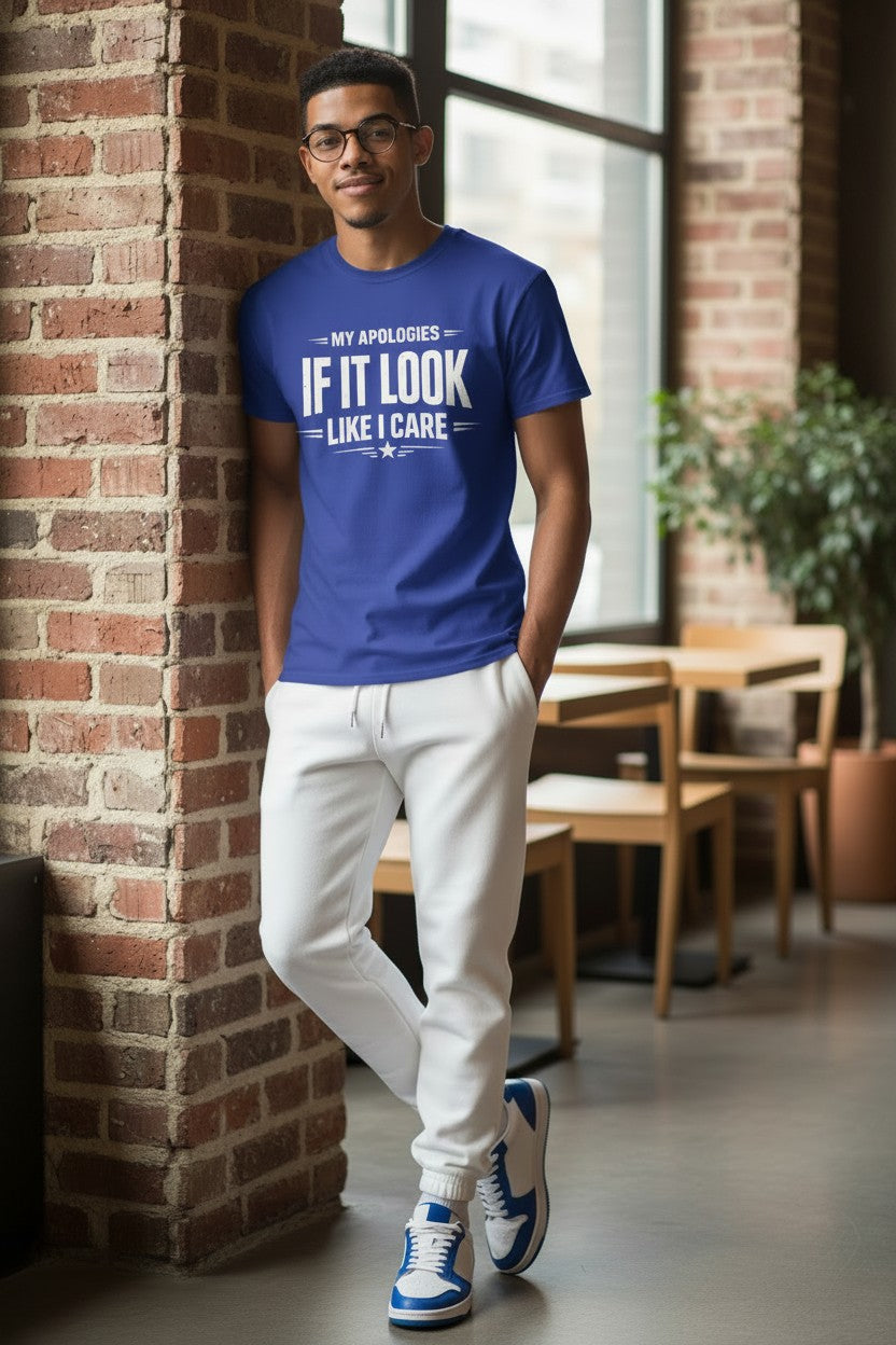 A man wearing a royal blue graphic t-shirt with the text 'MY APOLIGIES IF IT LOOK LIKE I CARE' in white, paired with white sweatpants and blue-and-white sneakers, leaning against a brick pillar in a modern indoor setting.