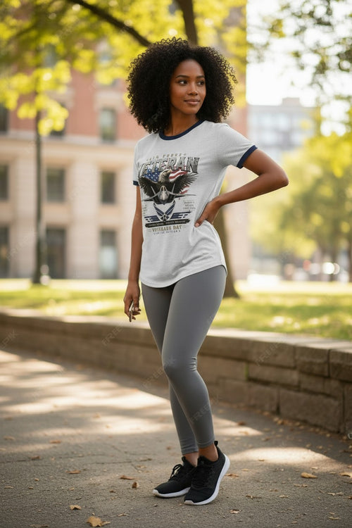 A woman stands outdoors on a paved path wearing a white ringer T-shirt with dark navy trim and a large eagle graphic on the front, paired with gray leggings and black sneakers. Trees and a building are blurred in the background.