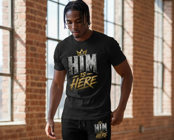 A man wearing a black graphic t-shirt with a distressed white and gold 'HIM IS HERE' print, standing in front of a brick wall with large windows.