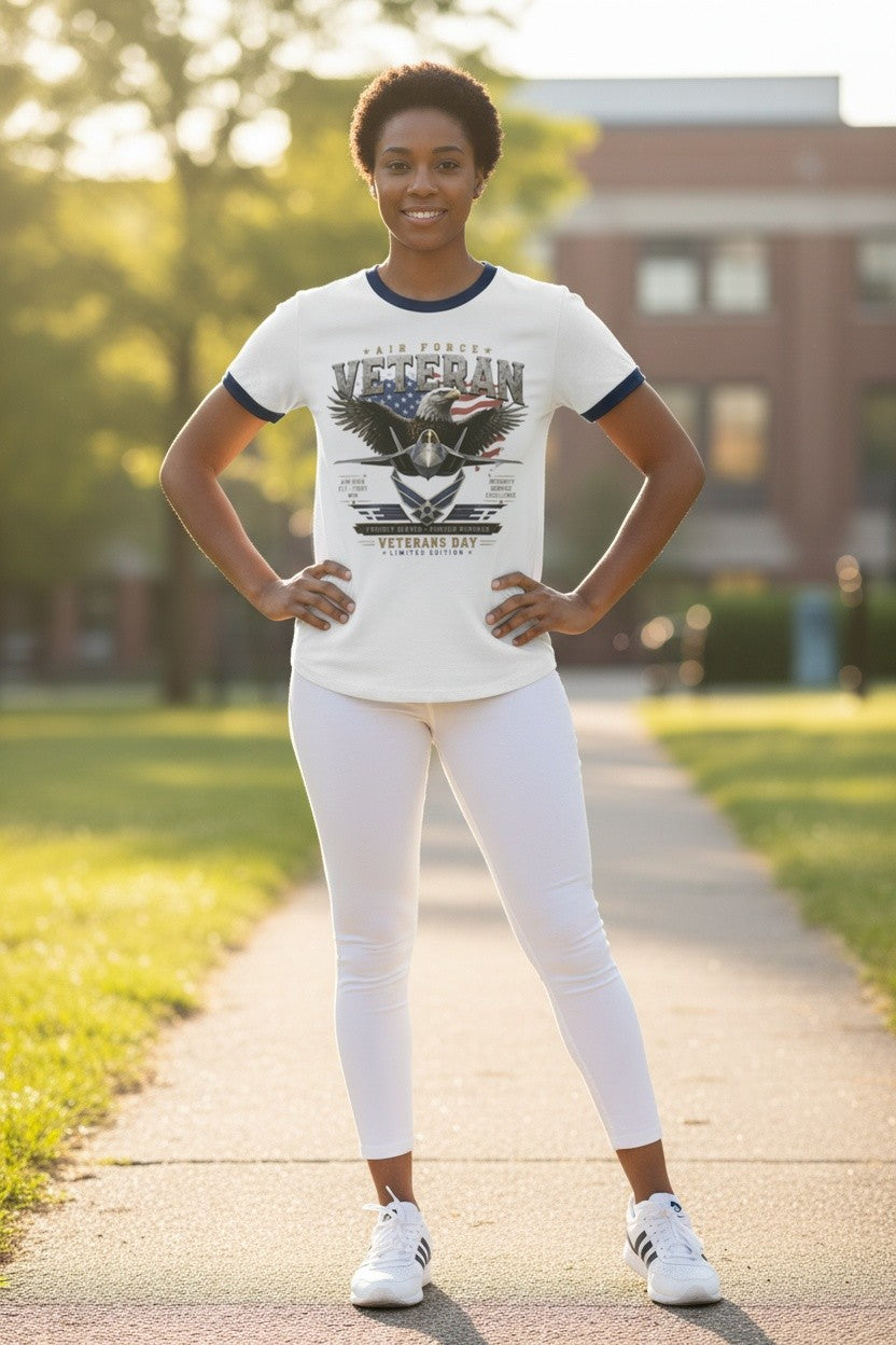 A woman stands on a sunlit pathway outdoors wearing a white ringer T-shirt with navy trim and a large patriotic veteran graphic, paired with white leggings and white sneakers.