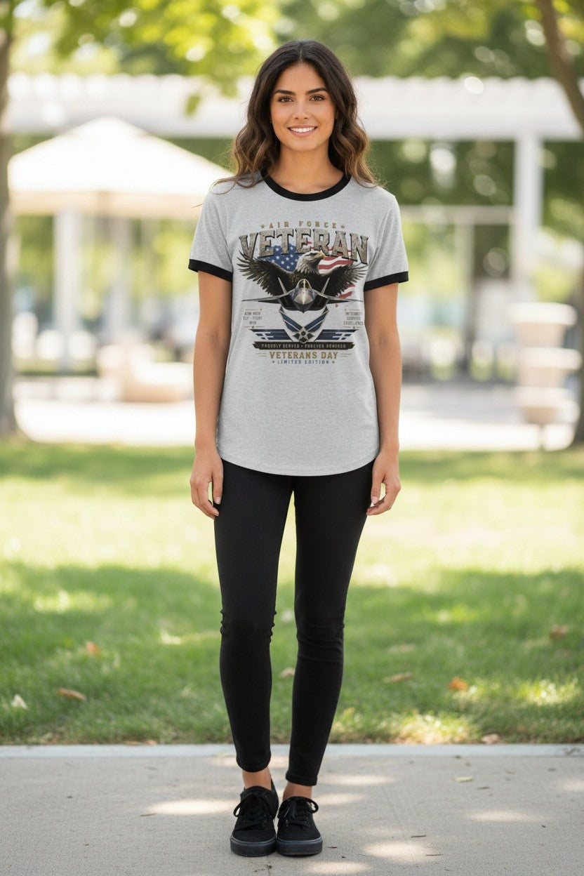 A woman stands outdoors on a paved path wearing a light gray ringer T-shirt with black trim and a large patriotic eagle graphic on the front, paired with black leggings and black sneakers.
