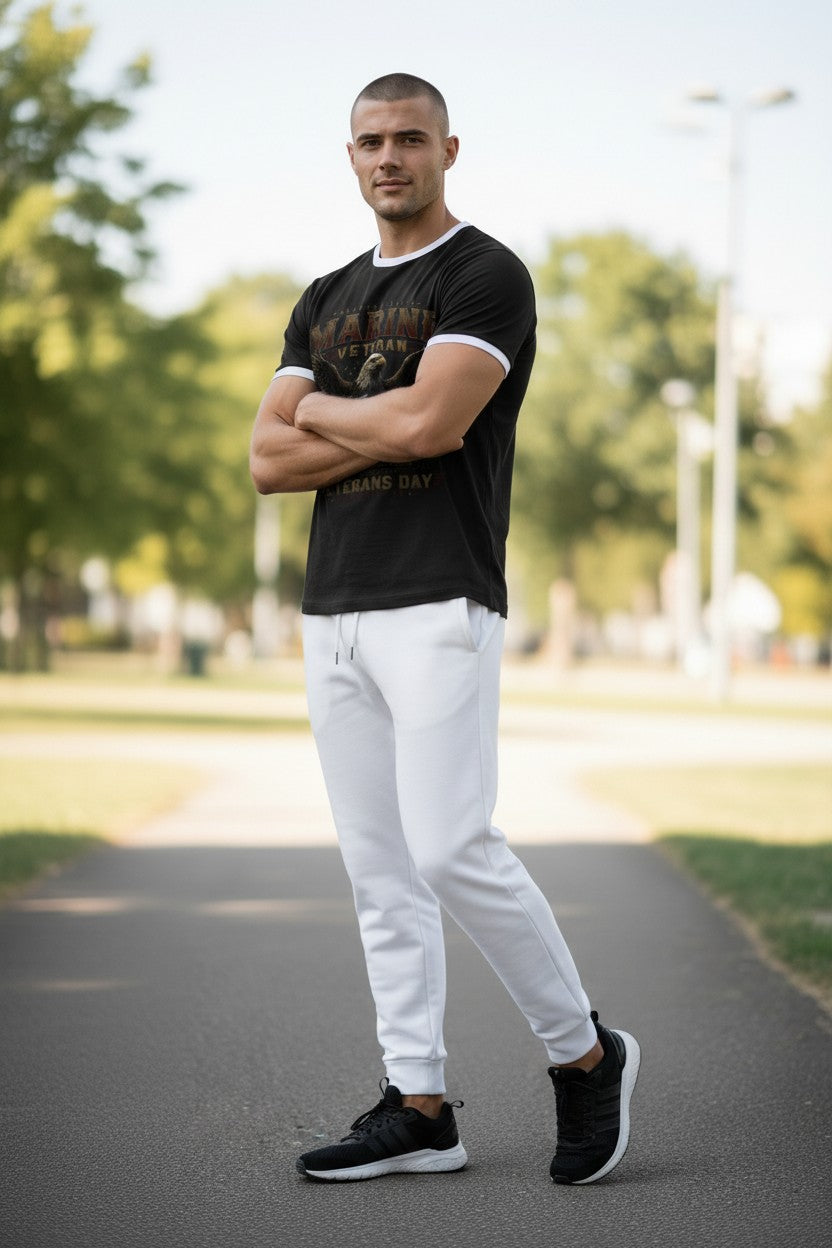 A man standing outdoors on a paved path wearing a black graphic t-shirt with white trim, white sweatpants, and black sneakers.