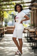 A woman modeling a white short-sleeve graphic T-shirt and matching white biker shorts in an outdoor patio setting with tables and greenery in the background.