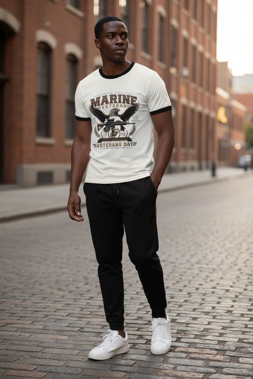 A man standing on a cobblestone street wearing a white ringer t-shirt with a black graphic print that reads 'MARINE VETERANS DAY', paired with black sweatpants and white sneakers.