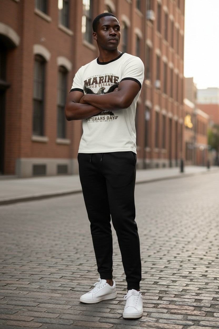 A man standing on a cobblestone street in front of brick buildings, wearing a white graphic t-shirt with black trim, black jogger pants, and white sneakers.