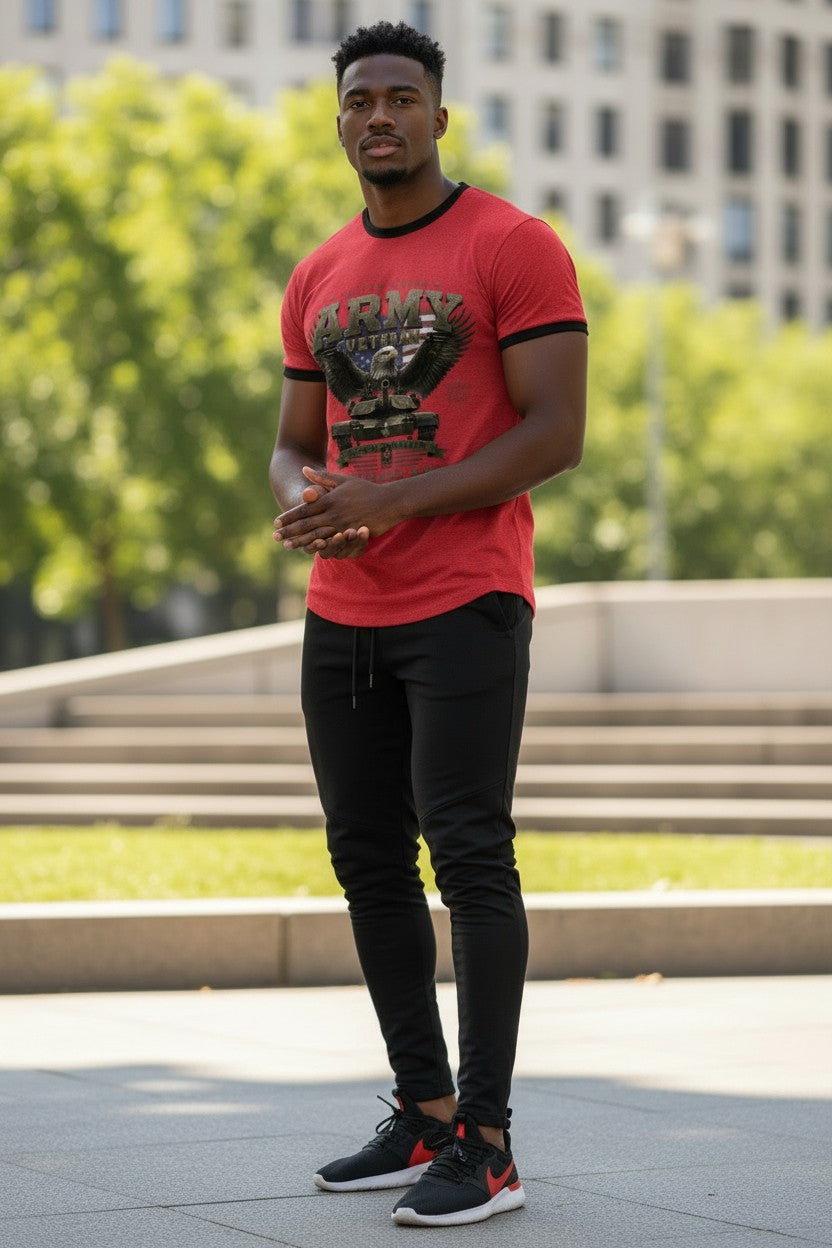 A man standing outdoors in a red graphic t-shirt with black trim, black pants, and black sneakers with red accents, set against a blurred background of green trees and a building.