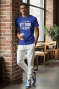 A man wearing a royal blue graphic t-shirt with the text 'MY APOLOGIES IF IT LOOK LIKE I CARE' in white, paired with white sweatpants and blue-and-white sneakers, leaning against a brick wall in a cafe setting.