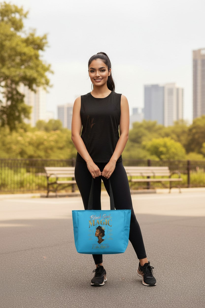 A woman in a black sleeveless top and leggings stands outdoors holding a bright blue tote bag with a gold-toned 'FASHION' print. The background shows a blurred park with trees, benches, and city buildings.