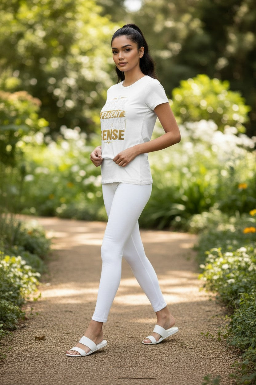 A woman walking on a garden path wearing a white athletic t-shirt with gold lettering that reads 'FIGHT DENSE' and matching white leggings.