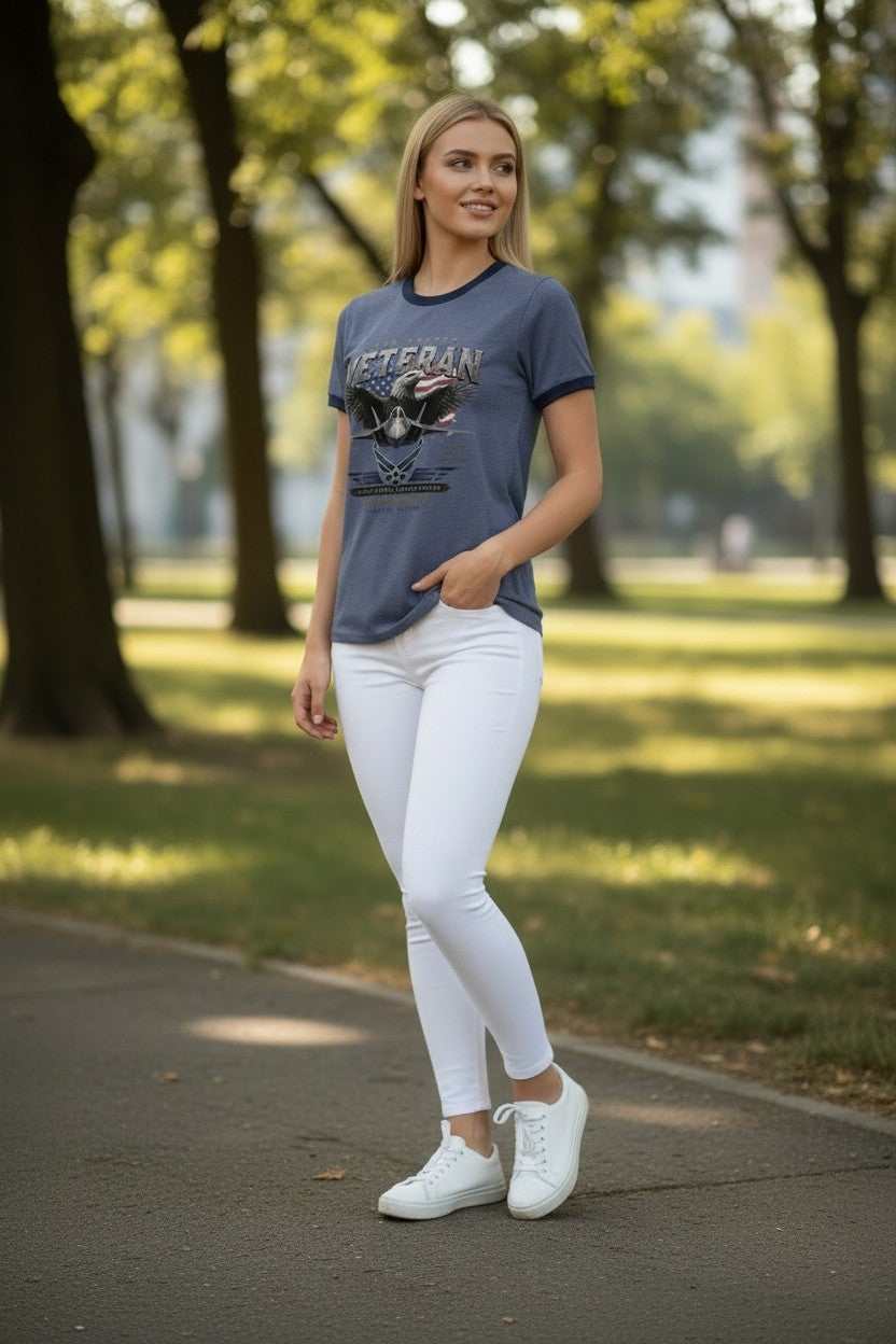 A woman standing on a paved path in a park, wearing a slate blue ringer t-shirt with a graphic print, white skinny pants, and white sneakers.