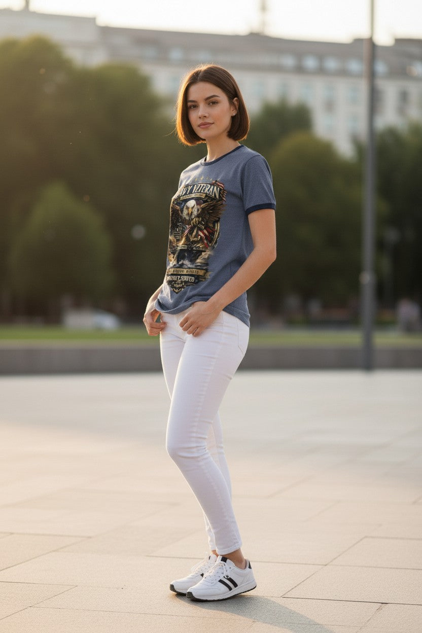 A woman stands outdoors wearing a slate blue graphic t-shirt, white skinny jeans, and white sneakers with black stripes. The background shows a softly blurred urban park setting with trees and a building.