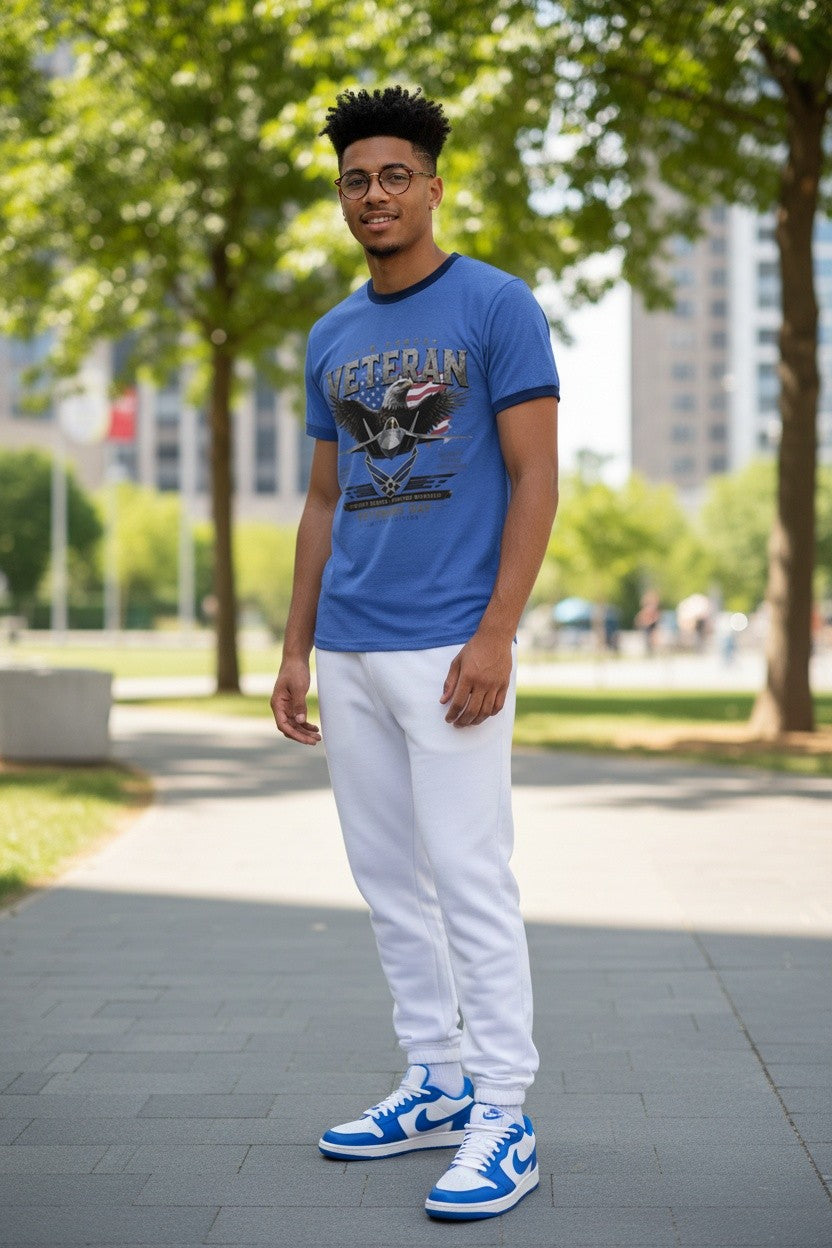 A man wearing a blue graphic t-shirt, white sweatpants, and blue and white sneakers stands on a paved path in a park with trees and buildings in the background.