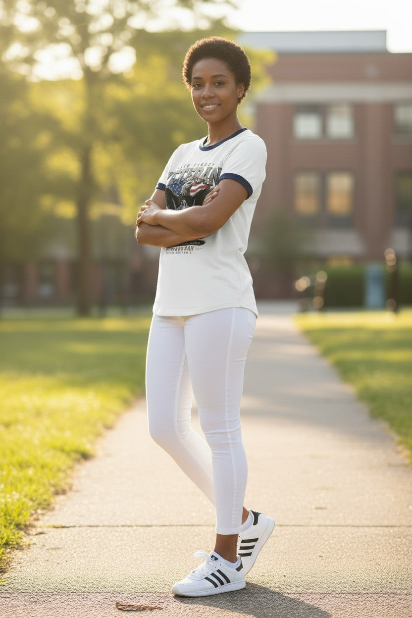 A woman stands on a sunlit outdoor path wearing a white graphic T-shirt with dark navy trim, white leggings, and white sneakers with black stripes. Trees and a brick building are blurred in the background.