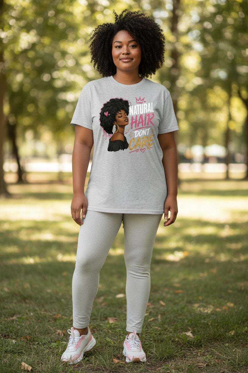 A woman standing outdoors on a grassy park path wearing a light gray graphic t-shirt, light gray leggings, and white sneakers. The t-shirt features a cartoon illustration of a Black woman with an afro and the text 'Natural Hair Don't Care' in pink and yellow.