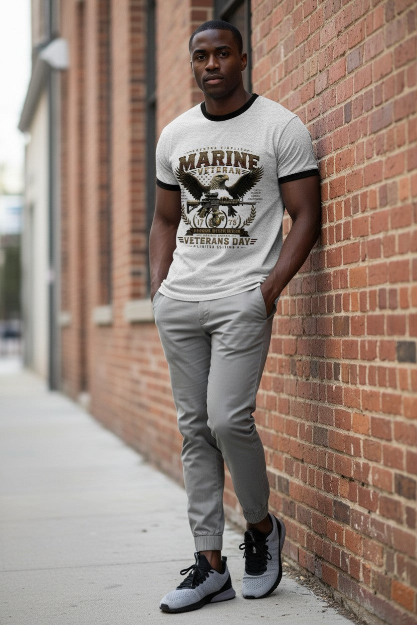 A man stands against a red brick wall wearing a white graphic t-shirt with black trim, gray jogger pants, and black and gray sneakers. The t-shirt features a dark graphic with the text 'MARINE' and 'VETERANS DAY'.