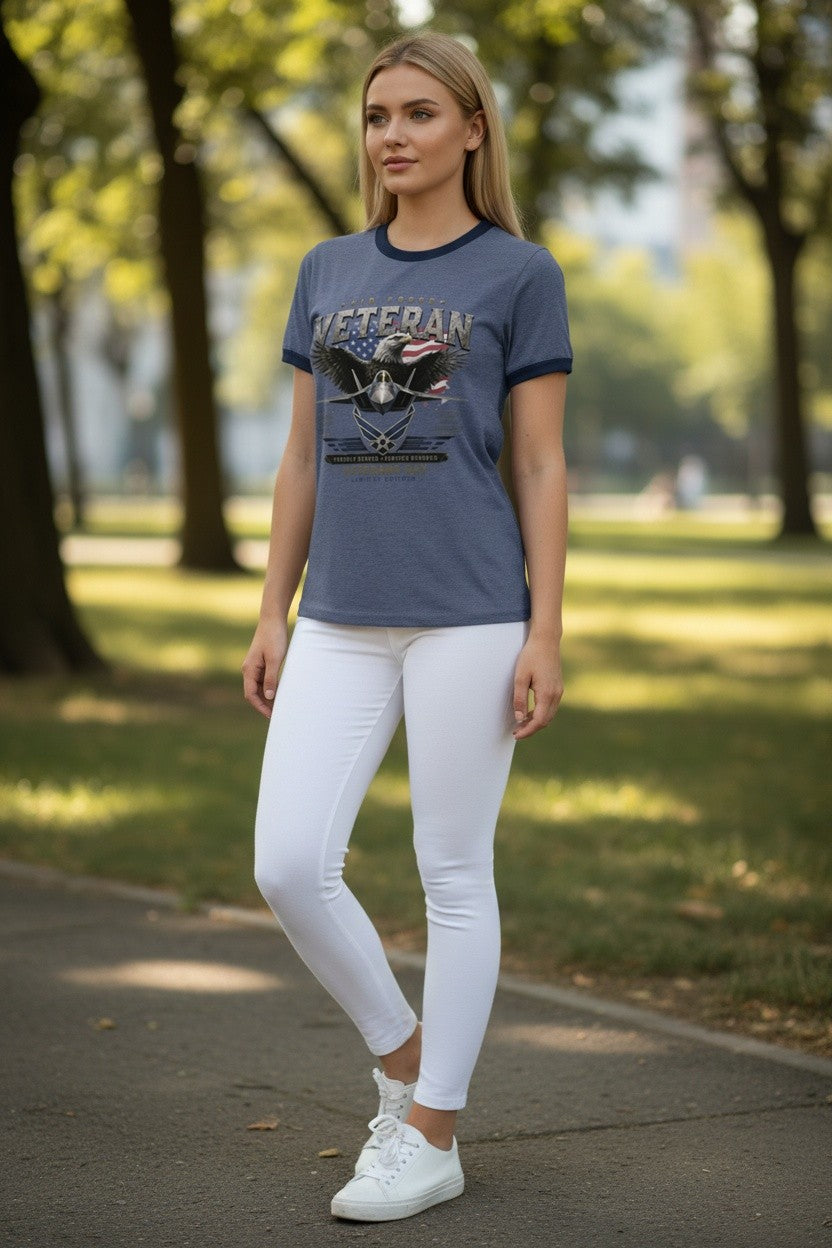 A woman standing on a paved path in a park, wearing a slate blue ringer t-shirt with a 'VETERAN' graphic, white skinny pants, and white sneakers.