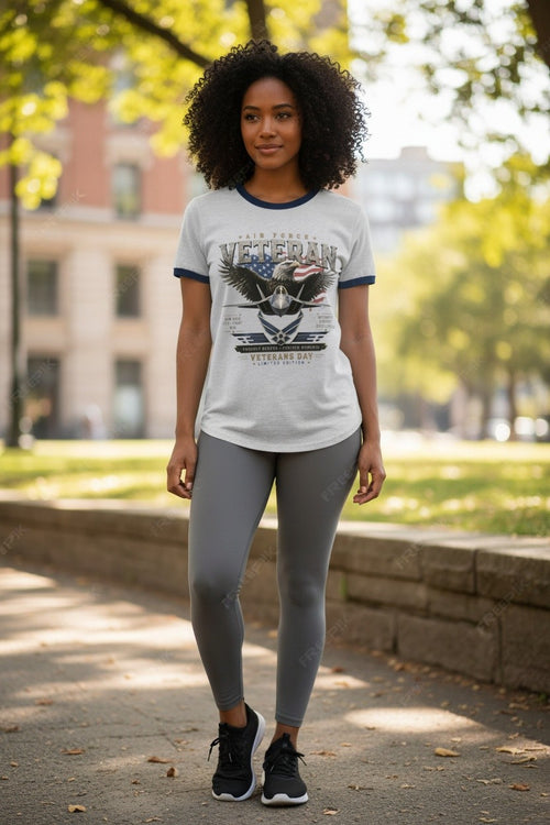 A woman stands on a sunlit park path wearing a white ringer T-shirt with navy trim and a large eagle graphic on the front, paired with gray leggings and black sneakers.