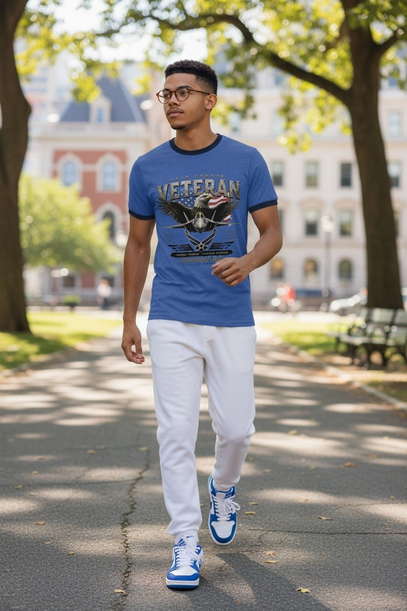 A man walking on a sunny park path wearing a blue graphic t-shirt with a 'VETERAN' eagle design, white sweatpants, and blue and white sneakers.