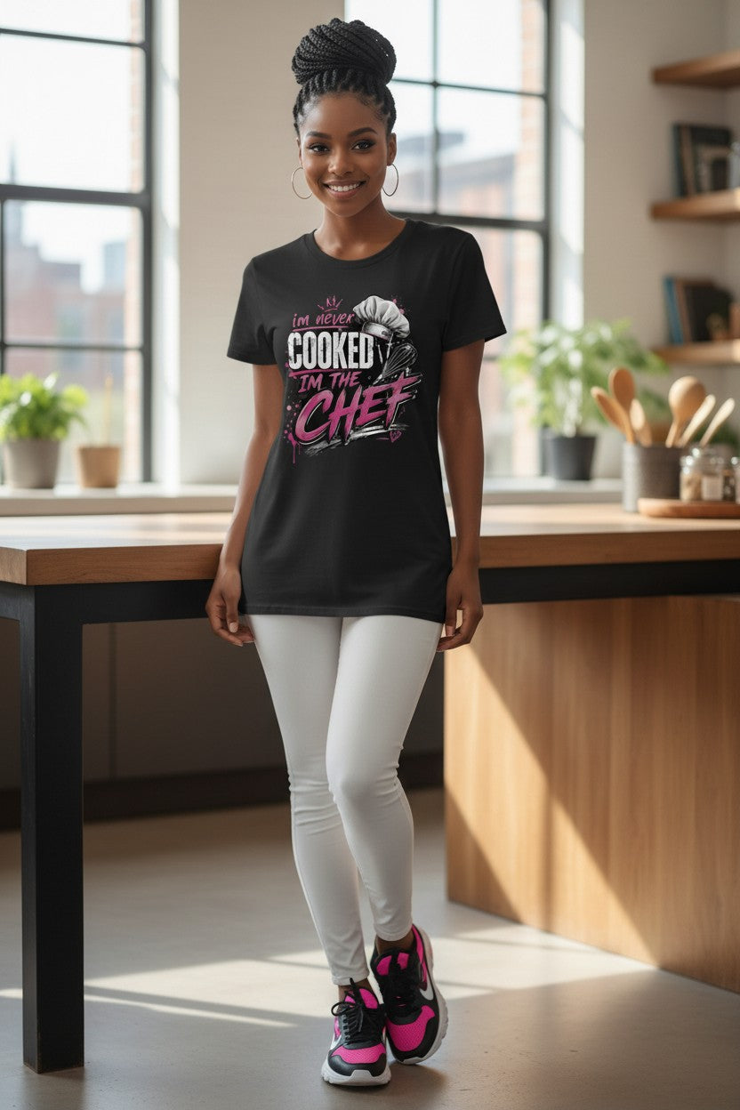 A woman stands in a bright kitchen setting wearing a black graphic T-shirt with the phrase 'I'M JUST COOKED UP THE CHEF' in white and pink lettering, paired with white leggings and pink-and-black sneakers.