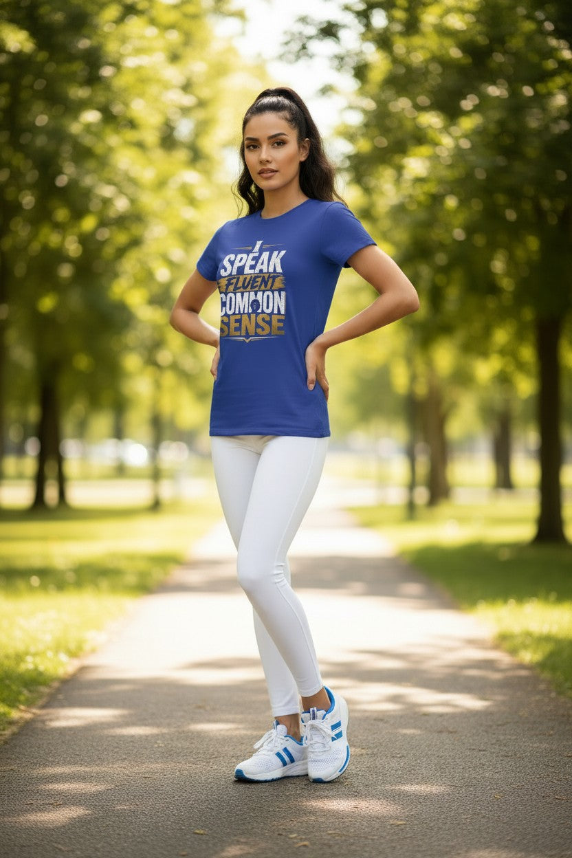 A woman stands on a tree-lined path wearing a royal blue graphic t-shirt with the text 'SPEAK LIKE A COMMON SENSE' in white and gold, paired with white leggings and white athletic sneakers with blue accents.