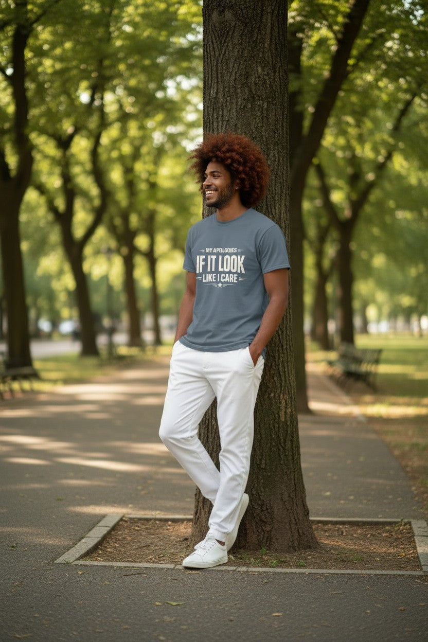 A man with curly brown hair leans against a tree on a park path, wearing a slate blue graphic t-shirt that reads 'IF IT LOOK LIKE CAFE' in white text, paired with white pants and white sneakers.