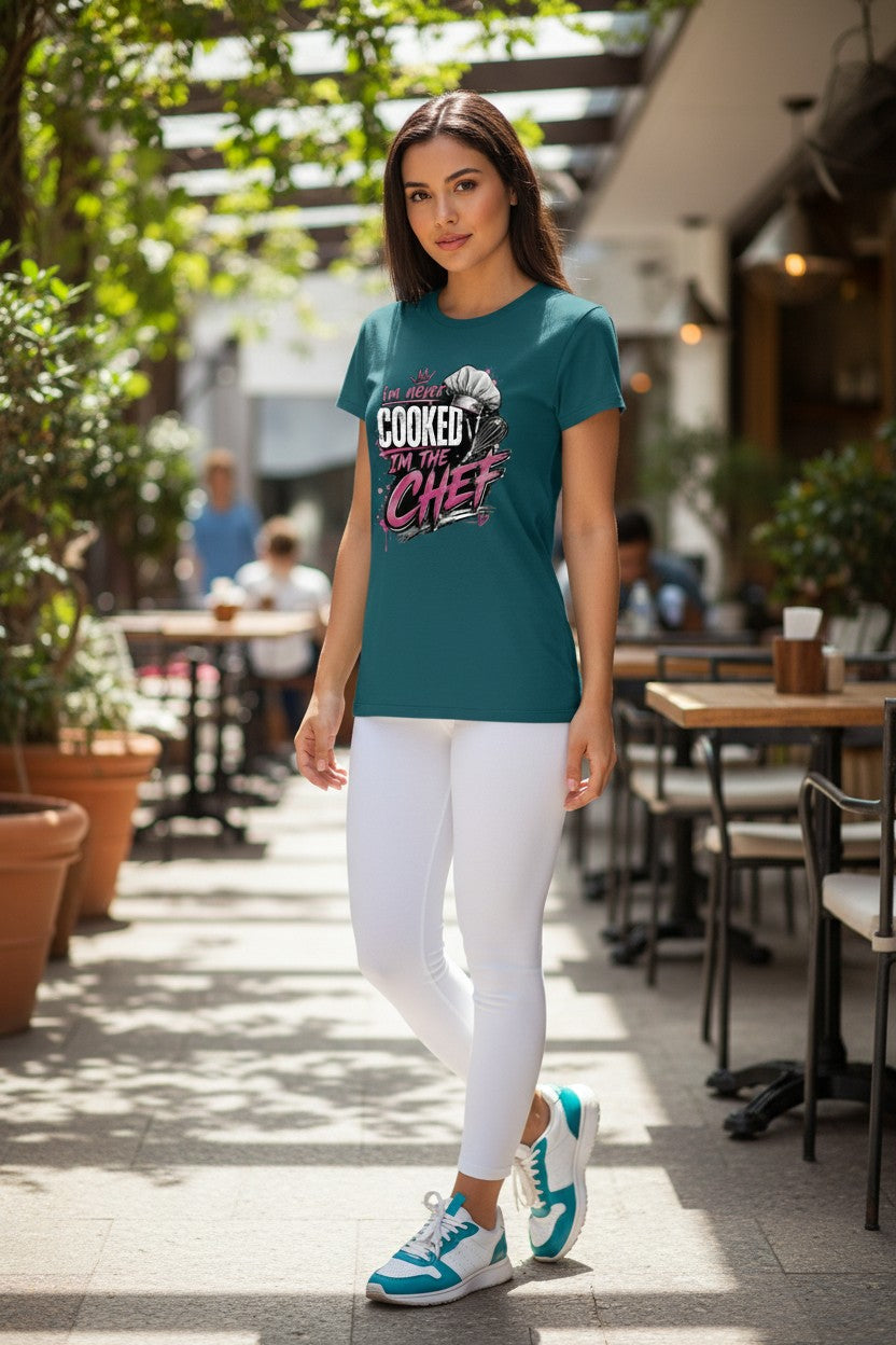 A woman stands in a sunlit outdoor cafe walkway wearing a teal graphic T-shirt with the words 'COOKED LIKE A CHEF' in white and pink, paired with white leggings and teal-and-white sneakers.
