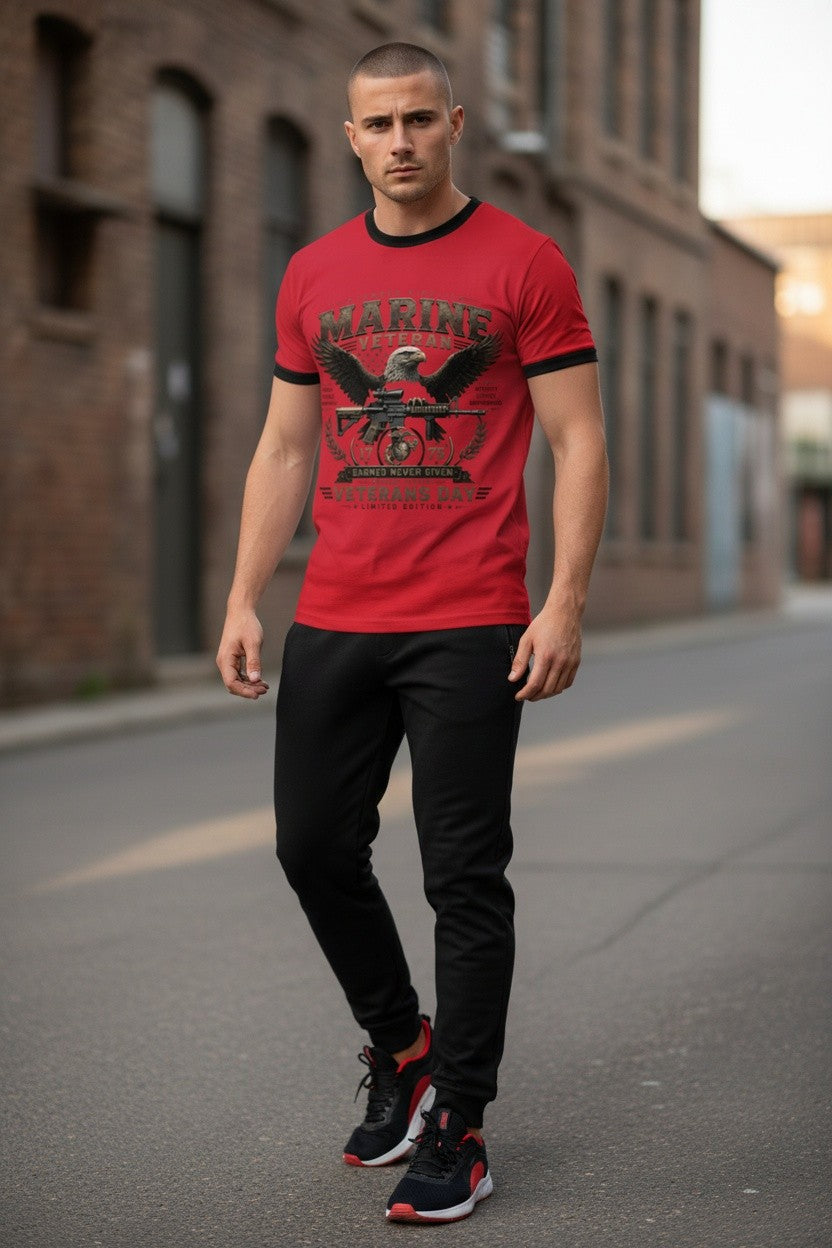 A man walking down an urban street wearing a red t-shirt with a black ringer collar and a distressed US Marine Corps eagle graphic on the front, paired with black pants and black and red sneakers.