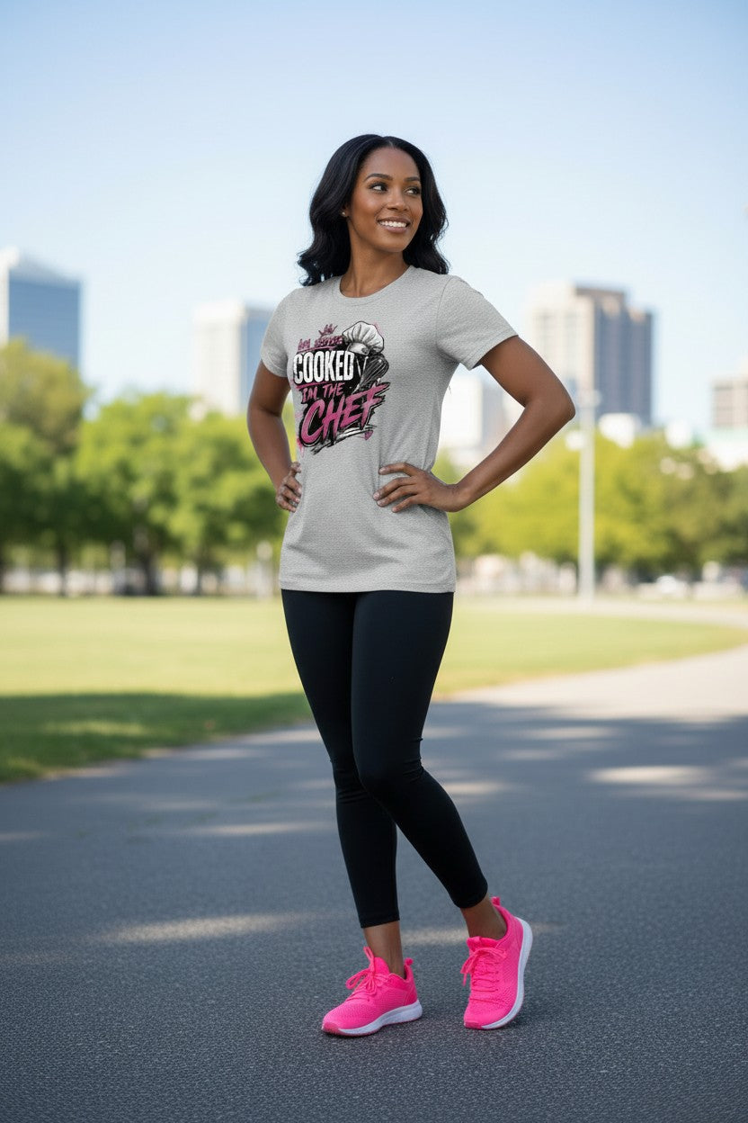 A woman posing outdoors on a paved path wearing a light gray graphic t-shirt with 'The Choked Chef' printed on the front, black leggings, and bright pink sneakers.