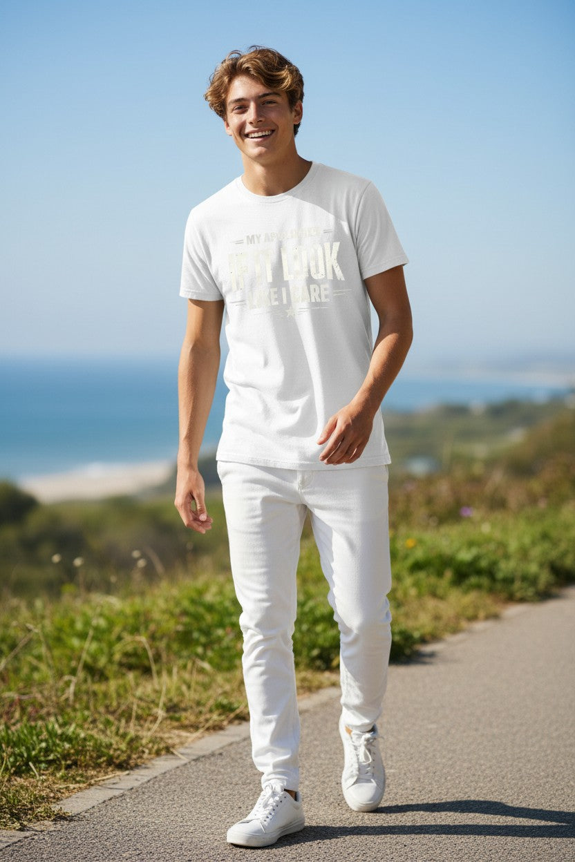 A smiling young man walking outdoors on a paved path wearing a white graphic t-shirt, white jeans, and white sneakers, with a blurred coastal landscape in the background.