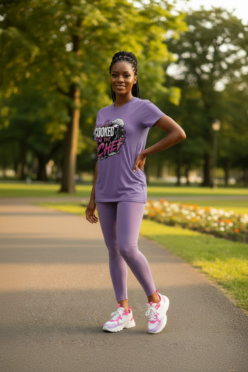 A woman stands on a paved path in a park wearing a lavender athletic t-shirt with a 'COOKED CHEF' graphic, matching lavender leggings, and pink and white sneakers.