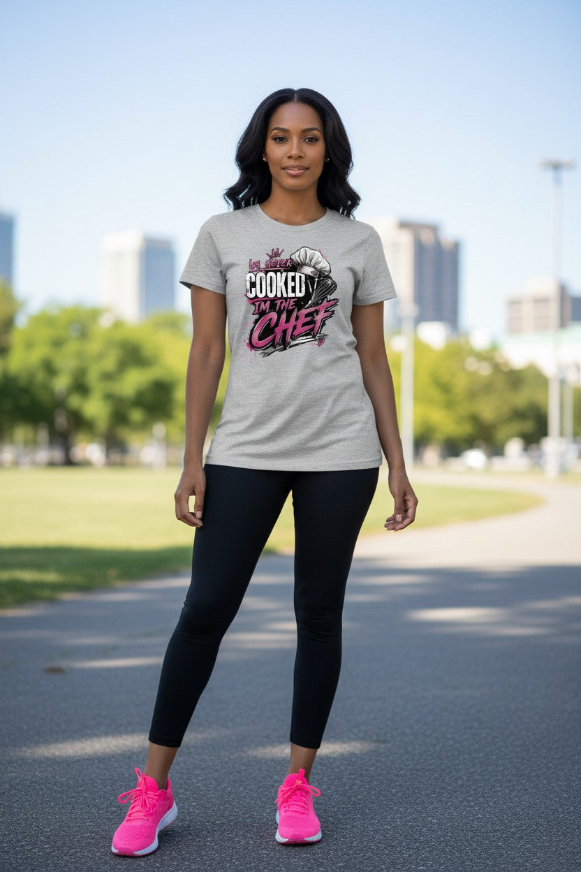 A woman standing outdoors on a paved path wearing a light gray graphic t-shirt with the text 'I'M COOKED AS CHEF' in pink and black, paired with black leggings and bright pink sneakers.