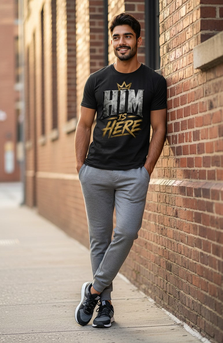 Man wearing a black t-shirt with 'HIM IS HERE' text, standing against a brick wall.
