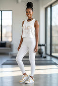 A woman modeling a white sleeveless crop top with 'CLASSY VIBE' printed in gold and silver, paired with white leggings and white sneakers with gold accents, in a bright modern indoor space.