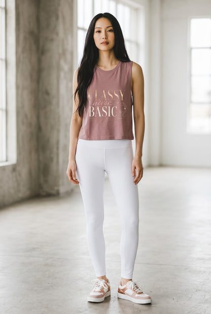 A woman stands in a bright, modern studio wearing a dusty rose sleeveless crop top with gold text that reads 'CLASSY is the BASIC?', paired with white leggings and white sneakers with gold accents.