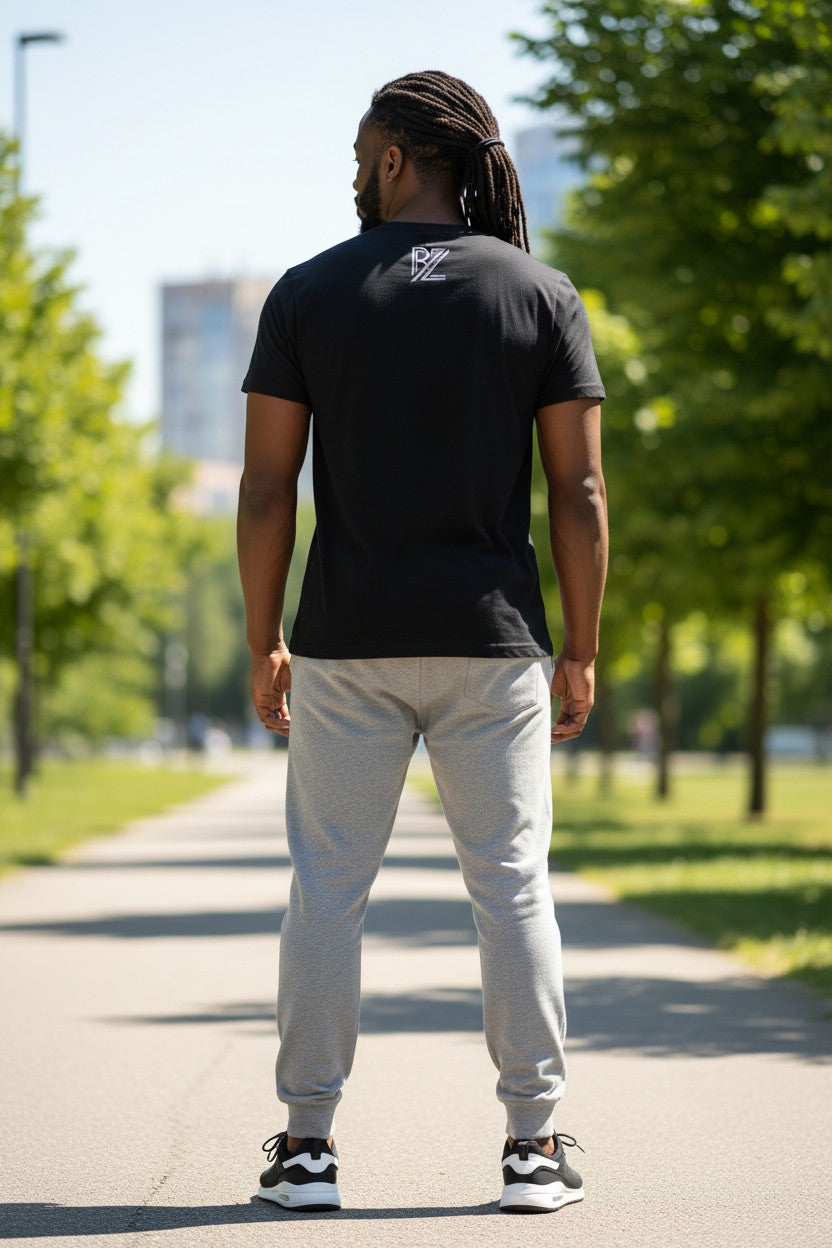 Person wearing a black t-shirt with a logo and light gray pants walking on a path with trees and buildings in the background.