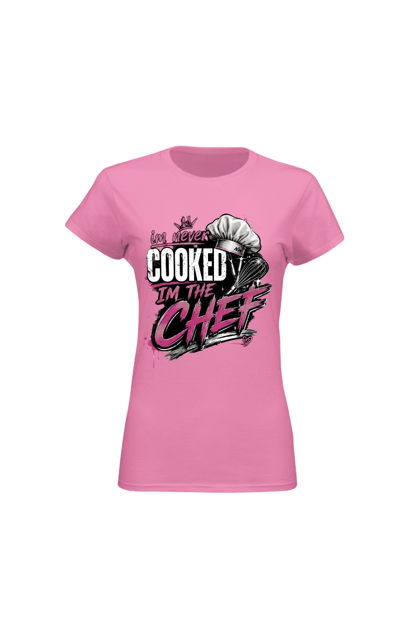 A pink short-sleeve crewneck t-shirt with a graphic on the front that reads 'I've never COOKED I'M THE CHEF' in stylized white, black, and pink lettering, accompanied by a chef's hat illustration.