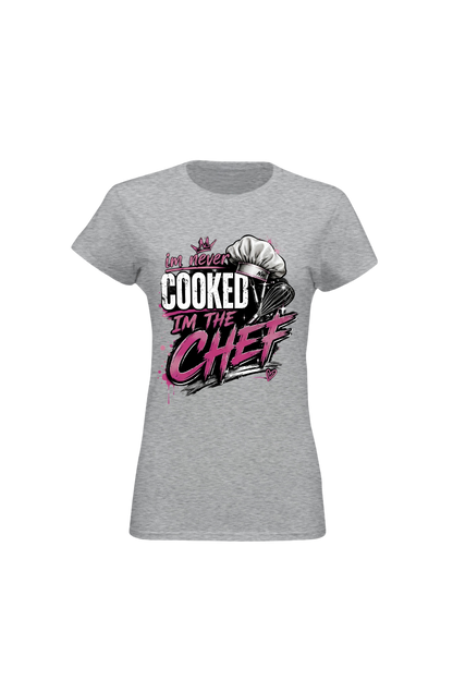 A women's heather gray short-sleeve t-shirt with a graphic on the front that reads 'I've never COOKED I'M THE CHEF' in white and pink lettering, accompanied by a chef's hat illustration.