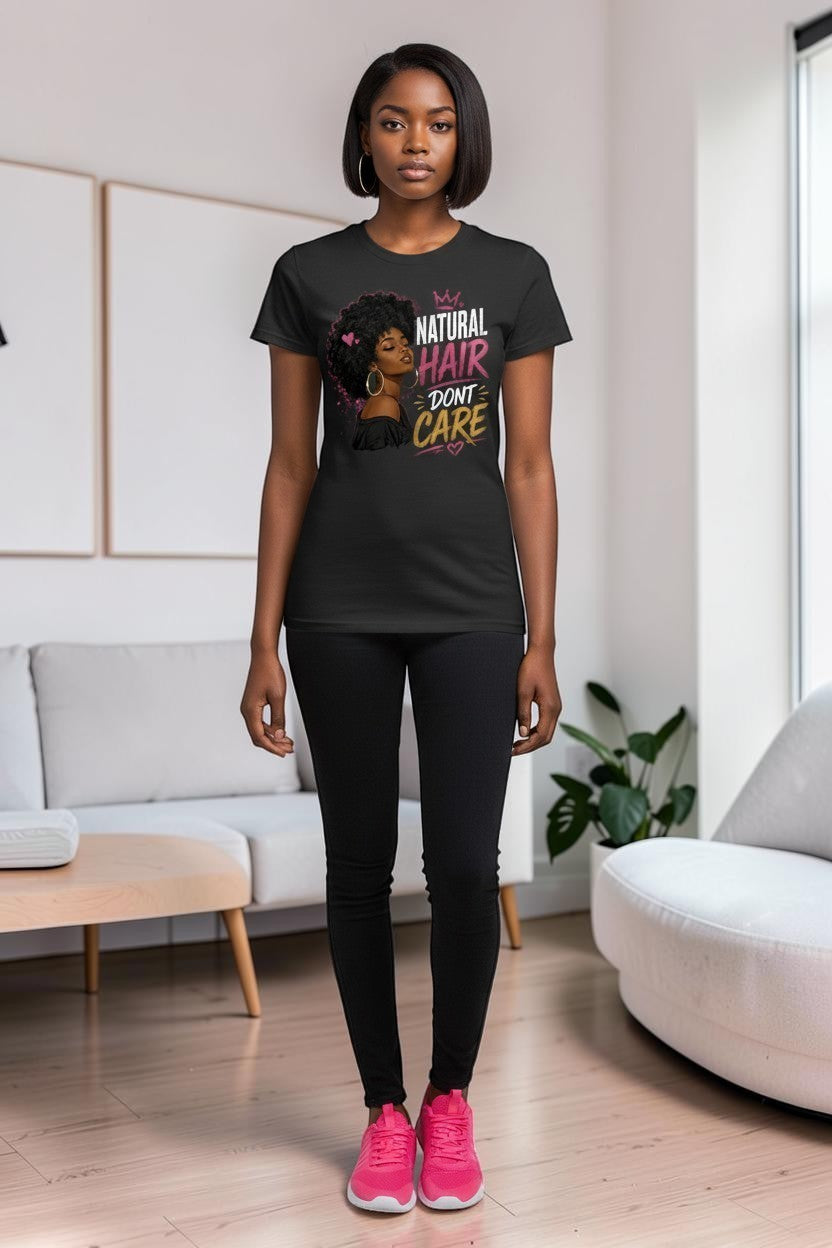 A woman modeling a black graphic t-shirt in a bright, modern living room. The t-shirt features a graphic of a Black woman with natural hair and the text 'YOUR NATURAL HAIR DON'T CARE'.