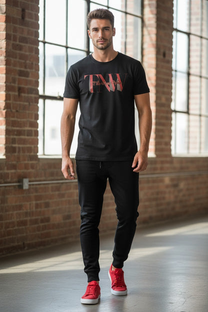 Man wearing a black t-shirt with red text, black pants, and red shoes in a room with large windows and brick walls.
