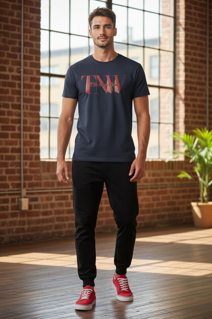 Man wearing a navy t-shirt with red text, black pants, and red sneakers standing in a room with large windows and brick walls.