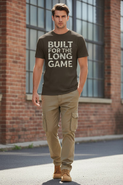 Man wearing a brown t-shirt with 'BUILT FOR THE LONG GAME' text, standing in front of a brick building.